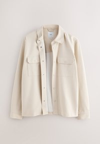 Veste chemise beige à manches longues avec boutons-pression, deux poches avant à rabat et un petit col, suspendue sur un fond uni.