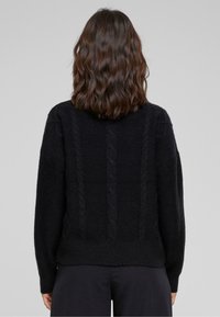 Schouderlang golvend bruin haar zichtbaar op een persoon die een zwarte kabelgebreide trui en donkere broek draagt, staande tegen een effen grijze achtergrond.