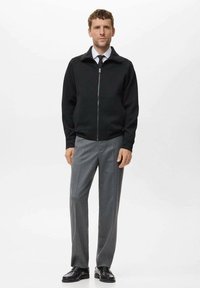 Chaqueta negra con cremallera y cuello alto, combinada con pantalones grises ajustados y zapatos de vestir negros. Diseño simple y corte slim.
