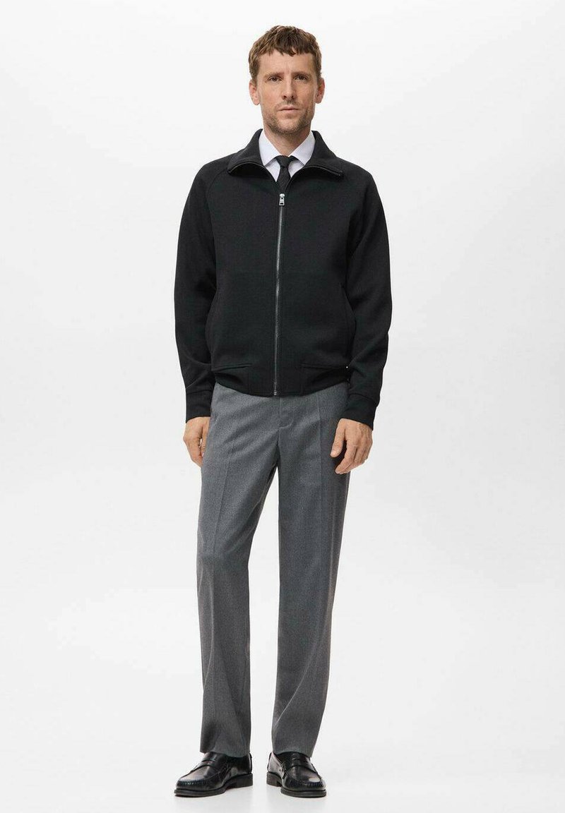 Schwarz zip-up Jacke mit hohem Kragen, kombiniert mit grauen maßgeschneiderten Hosen und schwarzen Businessschuhen. Einfache Gestaltung und schlanker Schnitt.