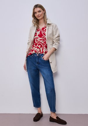 Kobieta w beżowej kurtce na czerwono-beżowej wzorzystej bluzce, niebieskich dżinsach i ciemnobrązowych mokasynach, stojąca na tle jasnoszarej ściany.