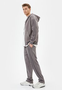 Sudadera gris con un bolsillo frontal y rayas en contraste; pantalones de chándal grises a juego con rayas laterales; combinados con zapatillas deportivas blancas.