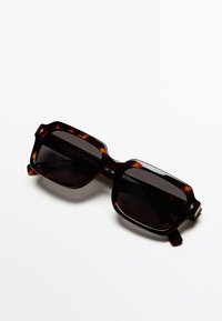 Zonnebril met schildpaddenkleurige montuur in een hexagonale vorm, donkere glazen en dikke randen met subtiele accenten aan de zijkanten.