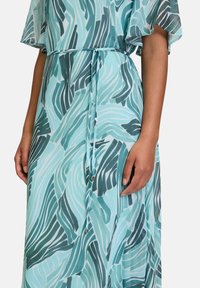 Leichtes, fließendes Kleid mit einem türkisfarbenen und dunkelgrünen Wellenmuster, das kurze Ärmel und einen Taillengurt mit metallischen Details aufweist.