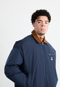 Temno modra zip-up jakna s ovratnikom iz korduroja v rjavi barvi, sprednjim žepom na zadrgo in logotipom na levem prsnem delu. Gladka, teksturirana tkanina.