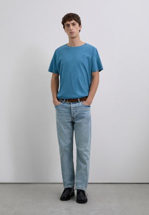 Joven de pie con las manos en los bolsillos de unos jeans azul claro, vistiendo una camiseta azul, cinturón marrón y zapatos negros sobre un fondo liso.