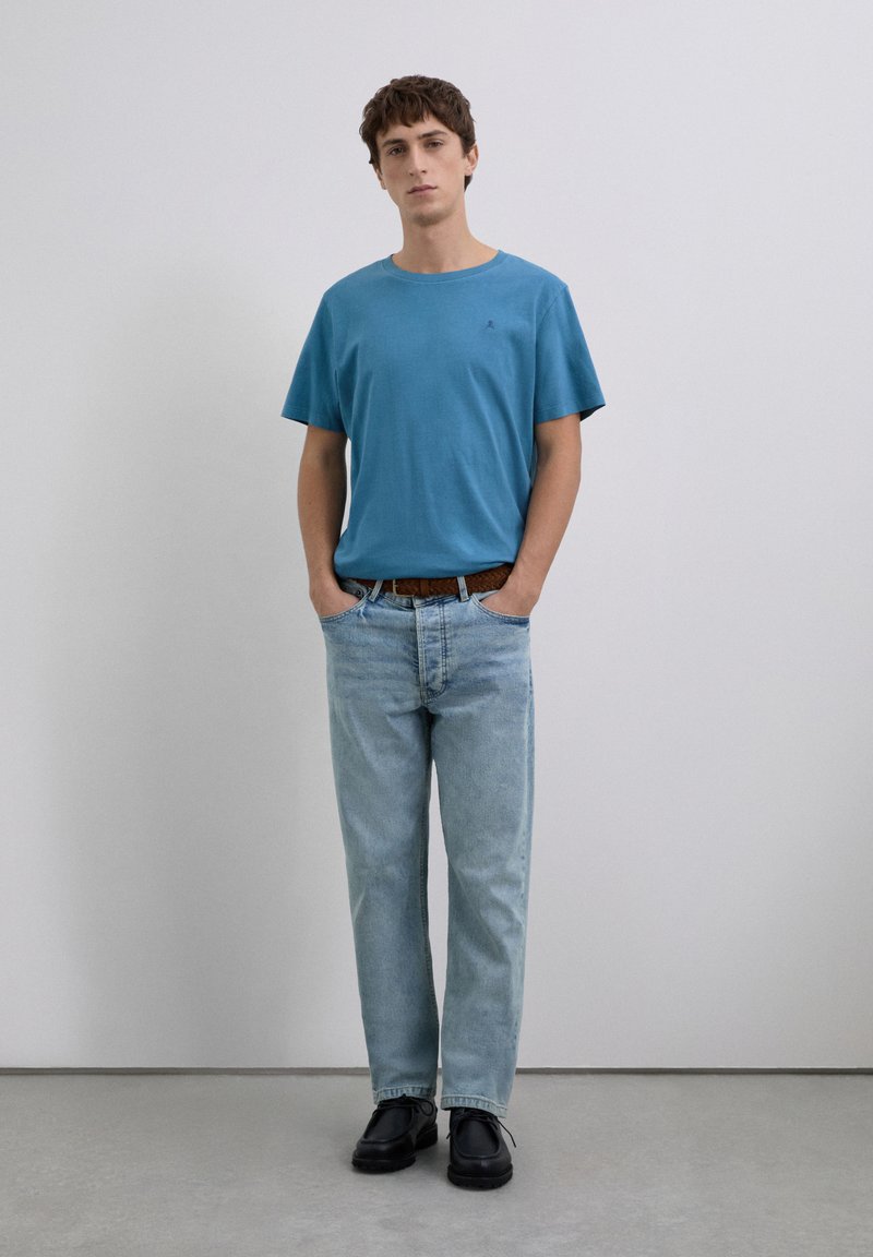 Ung mand står med hænderne i lommen på lyseblå jeans, iført en blå t-shirt, brun bælte og sorte sko mod en ensfarvet baggrund.