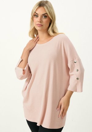 Lyshåret kvinde iført en løs, ribstrikket lyserød trøje med tre sølvknapper på ærmet, poserer med den ene hånd nær halsen.