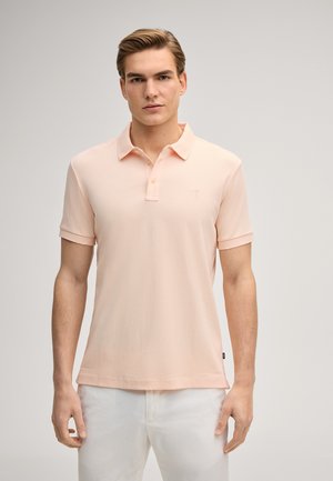 Giovane uomo in piedi, volto rivolto in avanti, che indossa una polo a maniche corte rosa chiaro e pantaloni bianchi su uno sfondo neutro.