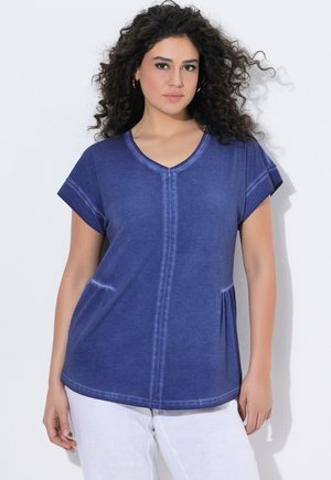 Frau mit lockigem Haar, die ein blaues kurzärmliges T-Shirt mit V-Ausschnitt und weiße Hose trägt, steht vor einem schlichten weißen Hintergrund.