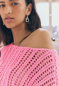 Top rosa lavorato a maglia, con spalle scoperte, caratterizzato da un motivo a trama aperta e morbida, che mette in risalto il filato testurizzato e esalta il tono della pelle. Orecchini circolari in argento.