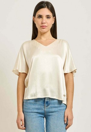Donna che indossa una blusa in raso color crema a maniche corte con scollo a V, abbinata a jeans azzurri, in piedi davanti a uno sfondo semplice.