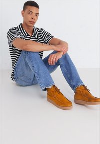 Botas de ante marrón hasta el tobillo con cordones, suelas de goma de color marrón claro. El modelo lleva una polo a rayas en blanco y negro y unos vaqueros azules, sentado.