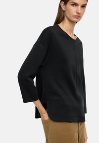 Czarna bluza z okrągłym dekoltem, rękawami do łokci i bocznymi kieszeniami. Gładka faktura, luźny krój i zakrzywiony dół.