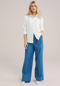 Camicia bianca a maniche lunghe con texture, abbinata a pantaloni jeans blu larghi. Il modello indossa scarpe bianche a punta e ha i capelli lunghi e lisci.