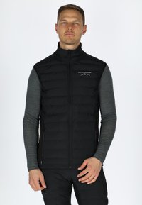 Schwarze isolierte Weste mit abgestepptem Design und Reißverschluss, mit hohem Kragen und Logodetail auf der Brust, getragen über einem grauen Langarmshirt.