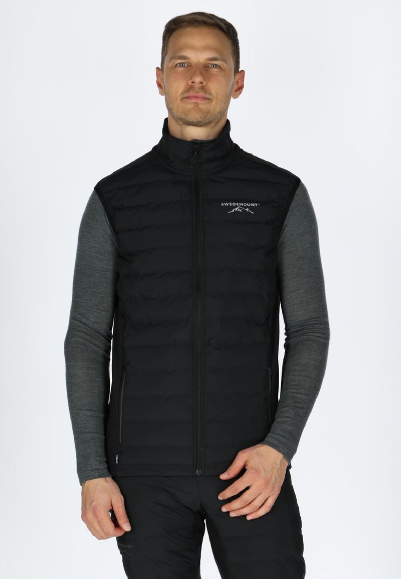 Schwarze isolierte Weste mit abgestepptem Design und Reißverschluss, mit hohem Kragen und Logodetail auf der Brust, getragen über einem grauen Langarmshirt.