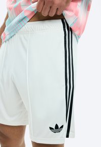 Άτομο που φοράει λευκό σορτς Adidas με μαύρες ρίγες στο πλάι και μαύρο λογότυπο Adidas κοντά στο τελείωμα, σηκώνοντας ένα πολύχρωμο πουκάμισο.