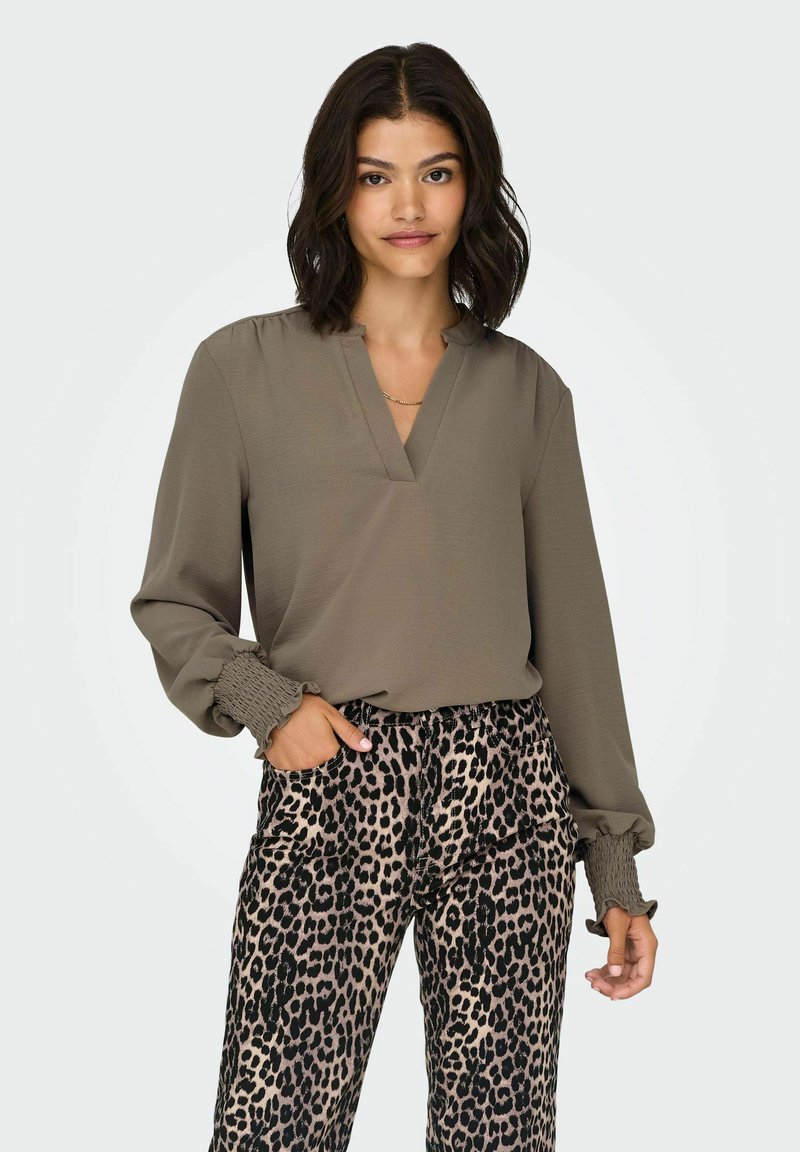Bluza dugih rukava, maslinaste boje, s izrezom na vratnom dijelu i volanima na rukavima, uparena s hlačama s leopard uzorkom u crnoj i bež boji.