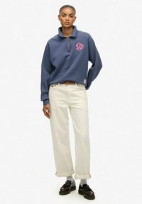 Kobieta stoi w niebieskiej bluzie z ćwierć zamkiem, białych szerokich spodniach nogawki z mankietami, szarych skarpetkach i czarnych mokasynach na tle jednorodnym.