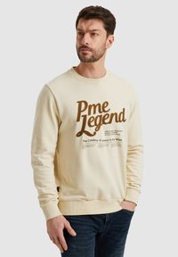 Sudadera color crema con cuello y puños acanalados. En el frente, presenta un gran lettering marrón que dice "Pme Legend" y un texto descriptivo debajo.