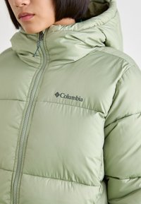 Jachetă puffer verde deschis cu glugă, prevăzută cu închidere cu fermoar, textură matlasată și logo-ul Columbia pe piept.