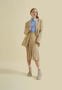 Beige oversize blazer över en ljusblå skjorta med knappar, parat med en veckad beige kjol och taupe plattformstövlar. Släta texturer genomgående.