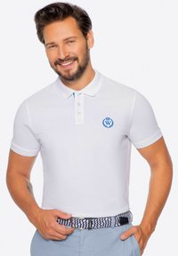 Biała koszulka polo o teksturowanym wykończeniu, z niebieskim haftowanym logo, zapięciem na trzy guziki i krótkimi rękawami. Połączona z wzorzystym paskiem.
