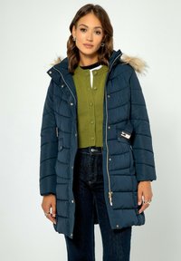Námořnická puffer bunda s kapucí podšitou umělou kožešinou, horizontálním prošíváním, bočními kapsami a zlatými kovovými doplňky. Nenošena přes zelený pletený kardigan.