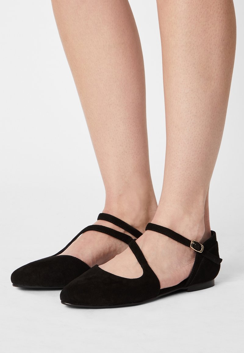 Svarta mocka ballerinas med spetsig tå, två ankelremmar och en liten guldklicka. Slät textur och minimalistisk design som visar foten.