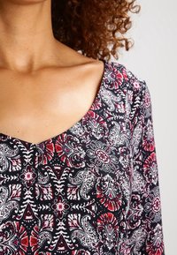 Tessuto nero a motivi con disegni floreali rossi, bianchi e grigi. Scollo a V e maniche corte con una finitura leggera e testurizzata.