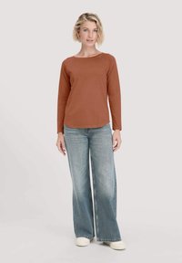 Rostfarbenslangärmliges Shirt mit einem Rundhalsausschnitt, kombiniert mit weit geschnittenen hellblauen Jeans und weißen Schuhen, vor einem neutralen Hintergrund stehend.