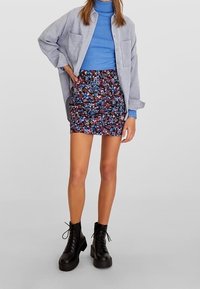 Lichtgrijs oversized shirt, blauwe turtleneck, minirok met bloemenpatroon en zwarte veterschoen met plateauzool. Soepele texturen en contrasterende kleuren.