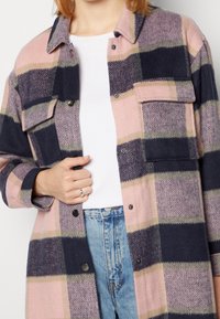 Osoba ubrana w różową, granatową i beżową koszulę w kratę z guzikami, nałożoną na białą koszulę i jasnoniebieskie dżinsy, trzyma rękę na kurtce blisko klatki piersiowej.