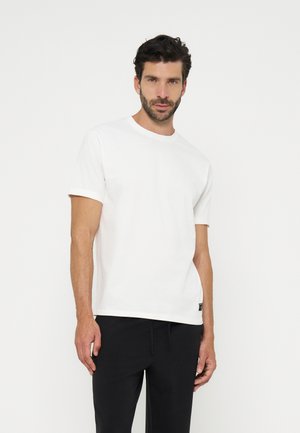 Homme aux cheveux courts et foncés avec une barbe, portant un t-shirt blanc uni à manches courtes et un pantalon noir, debout devant un fond blanc.