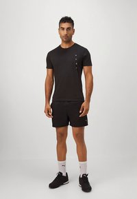 Camiseta preta de manga curta com um pequeno logótipo, combinada com calções pretos e sapatilhas pretas. O conjunto inclui meias desportivas brancas.