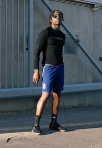 Top preto de manga comprida com design gráfico, calções azuis com logótipo da Itália, ténis pretos, em pé numa superfície pavimentada contra uma parede.