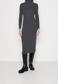 Donna che indossa un abito midi a maniche lunghe in maglia a coste grigio scuro con collo alto, abbinato a stivali neri con tacco largo fino al ginocchio, in piedi su un pavimento bianco.
