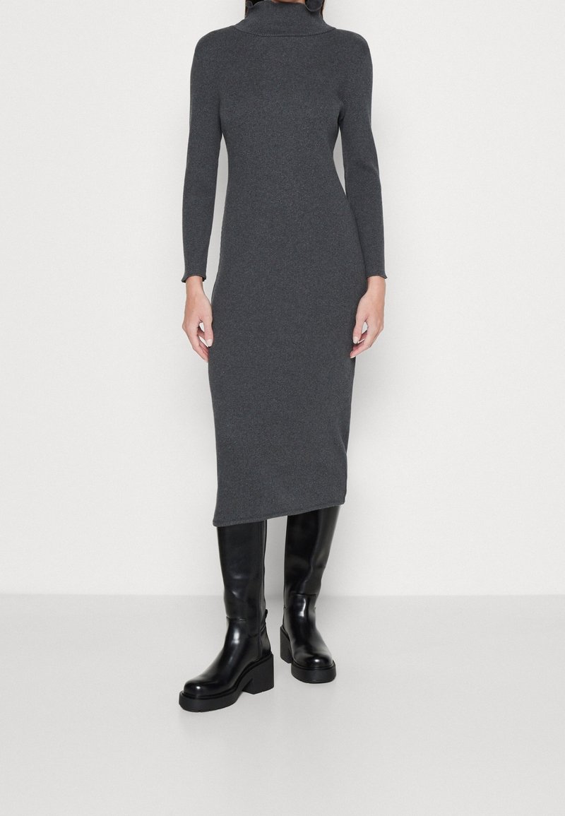 Donna che indossa un abito midi a maniche lunghe in maglia a coste grigio scuro con collo alto, abbinato a stivali neri con tacco largo fino al ginocchio, in piedi su un pavimento bianco.
