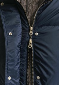Marineblauwe pufferjack met een rits aan de voorkant, gouden hardware en zilveren drukknoopjes; gevoerd met een textuur gebreide stof in een neutrale tint.