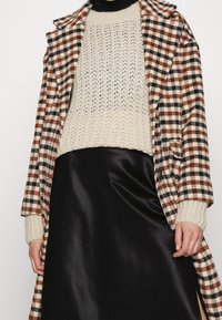 Braun- und schwarz karierter Mantel, der über einem cremefarbenen Strickpullover und einem schwarzen Satinrock getragen wird. Verfügt über gerippte Bündchen und ein tailliertes Design.