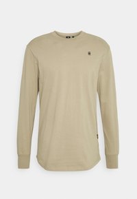 Maglietta beige a maniche lunghe con colletto girocollo, con piccolo logo nero "G RAW" sul petto e maniche con polsini, su sfondo bianco.