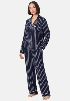 Femme debout pieds nus portant un pyjama rayé bleu marine à manches longues avec des boutons, une bordure blanche et une poche poitrine.