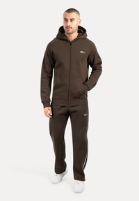 Sudadera con cremallera marrón, con bolsillos frontales y logo blanco, combinada con joggers marrones a juego que tienen rayas laterales blancas y zapatillas blancas.