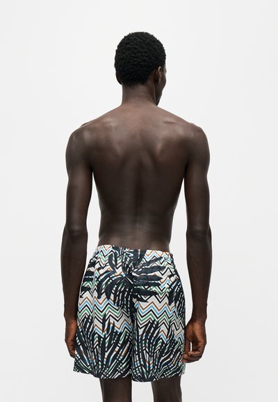 Homme à la peau foncée et aux cheveux courts et bouclés vu de dos portant un short de bain à motifs avec des dessins en zigzag et des feuilles.