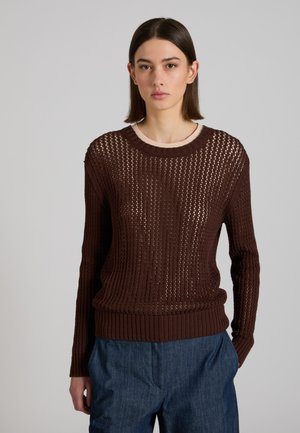 Jeune femme portant un pull brun à mailles ajourées par-dessus un haut beige et un pantalon ample bleu foncé, debout devant un fond gris uni.