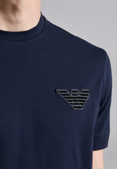 Navyblaues Crew-Neck T-Shirt aus strukturiertem Stoff mit einem geprägten schwarzen Logo auf der linken Brust, kurzen Ärmeln und einer taillierten Passform.
