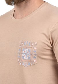 Kortermet beige t-skjorte med en mønstret lomme som har geometriske design i blå, oransje og kremfarger. Glatt tekstur.