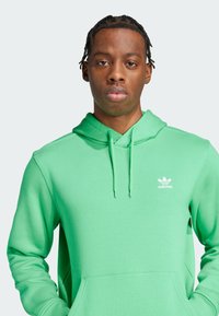 Groene hoodie van zacht materiaal, met een trekkoord capuchon, een voorzak en het witte Adidas-logo op de borst. Standaard pasvorm.