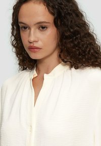 Crème-kleurige blouse van een gestructureerde stof, met een ontspannen kraag, knoopsluiting aan de voorkant en zachte plooien. Geschikt voor verschillende gelegenheden.